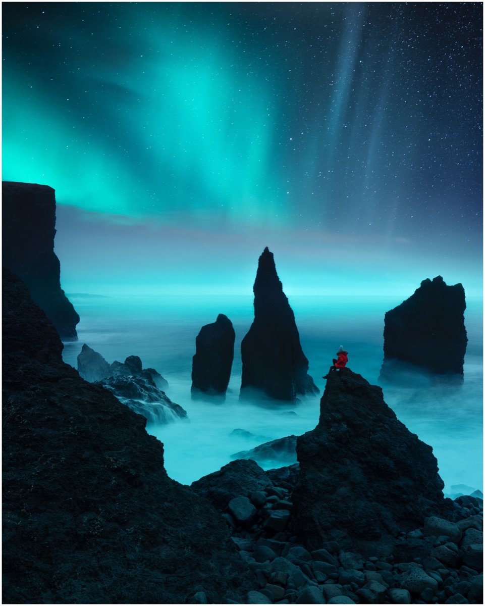 Green aurora over dark sea stacks, with a figure in red seated on a rock